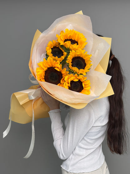 A person in a white long-sleeve top and light pants stands against a plain gray background, holding a bouquet of sunflowers wrapped in pale yellow tissue paper in front of their face.